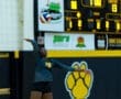 A volleyball player serves the ball during a game, with the scoreboard showing the score in the background.