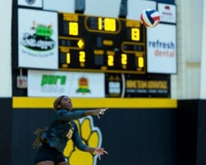 Volleyball player serving during a match, with the scoreboard showing a close game at 18-13.
