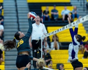 A volleyball player in action jumps to spike the ball over the net as a defender attempts to block. Energetic game moment.