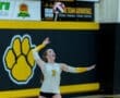 High school volleyball player serving a ball during a match, with a school logo in the background.