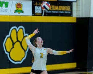 High school volleyball player serving a ball during a match, with a school logo in the background.