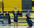 Volleyball player sets the ball during a game, with teammates and commentators visible in the background.