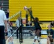 High school volleyball match action, featuring a player jumping to spike the ball over the net while teammates watch.