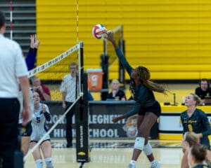 High school volleyball match action, featuring a player jumping to spike the ball over the net while teammates watch.