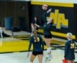 Volleyball player serves the ball during practice, with teammates observing in a gym with a black and yellow theme.