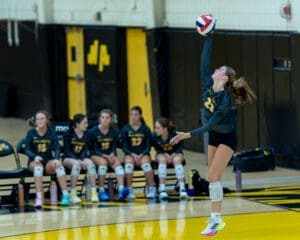 Volleyball player jumps to serve with teammates on the bench in the background during a game.