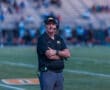 A football coach stands on the field with crossed arms, wearing a headset and team gear, focused on the game.