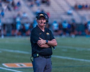 A football coach stands on the field with crossed arms, wearing a headset and team gear, focused on the game.