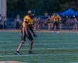 Football player in yellow uniform, ready in a defensive stance on the field during a game, with cheerleaders in the background.