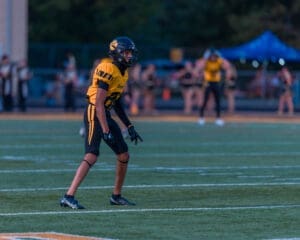Football player in yellow uniform, ready in a defensive stance on the field during a game, with cheerleaders in the background.