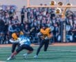 High school football game action: player 18 evades tackle by opponent 20 on the field with a marching band in the background.