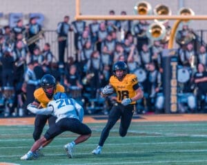 High school football game action: player 18 evades tackle by opponent 20 on the field with a marching band in the background.