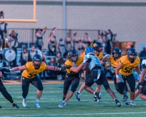 High school football players in yellow and black uniforms tackle an opponent on the field during a game at dusk.