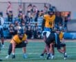 Football players in action during a game, with a quarterback in yellow jersey preparing to pass.