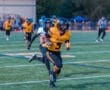 Player in yellow football uniform sprinting with the ball, evading a defender during a night game on a field.