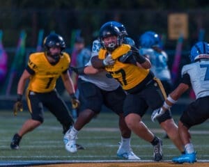 High school football players in yellow and blue jerseys compete during an evening game on the field.