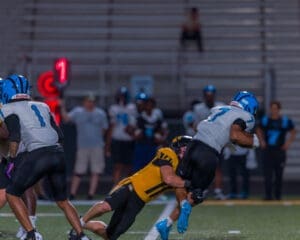 Football players in action during a night game, with a defender tackling the ball carrier on the field.