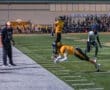 A football player in yellow dives for a catch near the sideline while a coach observes intensely.