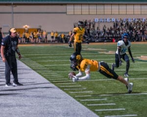 A football player in yellow dives for a catch near the sideline while a coach observes intensely.