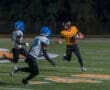 Player in yellow jersey dodges defenders in blue helmets during a night football game on a well-lit field.
