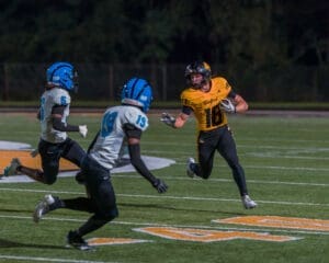 Player in yellow jersey dodges defenders in blue helmets during a night football game on a well-lit field.