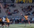 High school football game with a player in yellow throwing a pass while defenders pursue him on the field.