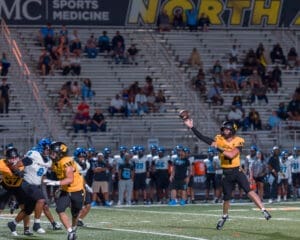 High school football game with a player in yellow throwing a pass while defenders pursue him on the field.