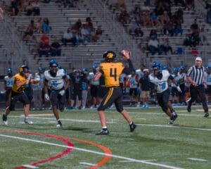 High school football game with a quarterback in yellow jersey throwing a pass while defenders in blue and black pursue him.