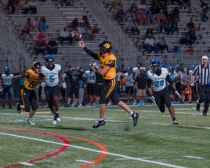 Quarterback in yellow jersey throws a football while being pursued by defenders in a night football game.
