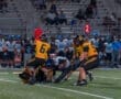 High school football game action, players in yellow jerseys tackling an opponent on the field under stadium lights.
