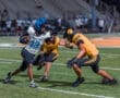 High school football players in action, one evading two defenders on the field under stadium lights.