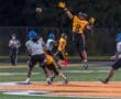 High school football player leaps to catch a pass during a night game on a well-lit field with players in action.
