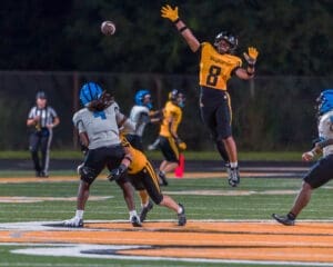 High school football player leaps to catch a pass during a night game on a well-lit field with players in action.