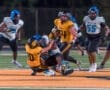 Two high school football players tackle each other on the field during a night game, surrounded by teammates.