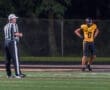 High school football player in yellow uniform stands on the field with a referee during a night game.