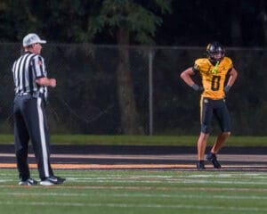 High school football player in yellow uniform stands on the field with a referee during a night game.