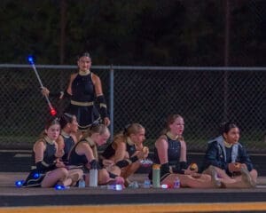 Drill team members sitting on the sidelines during a night performance, enjoying snacks and preparing for their routine.