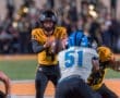 Quarterback in yellow jersey prepares to pass the football under pressure from a defender during a night game.