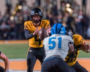 Quarterback in yellow jersey prepares to pass the football under pressure from a defender during a night game.
