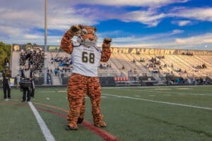 North Allegheny mascot in a tiger costume performing at a football game with fans and band visible in the background.