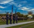 Color guard presenting flags at a high school football game, with a lively halftime show and scoreboard in the background.