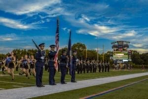 Color guard presenting flags at a high school football game, with a lively halftime show and scoreboard in the background.