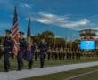 Color guard marching with the American flag at a football field, with a scoreboard and spectators in the background.