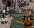 A band member in uniform stands beside a tiger mascot on a football field during a school event.
