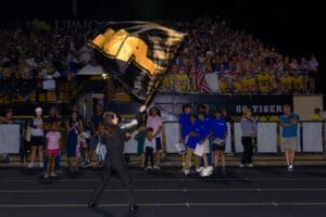 Cheerleader waving a black flag at a nighttime sports event, with an excited crowd in the background.