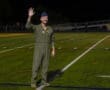Military officer in flight suit waving cheerfully on a sports field during a nighttime event.