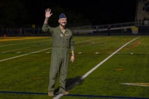 Military officer in flight suit waving cheerfully on a sports field during a nighttime event.