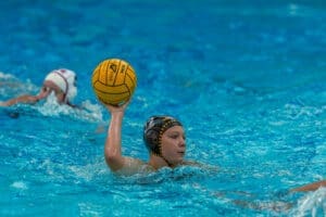 A female water polo player prepares to throw the ball while teammates swim in a blue pool.