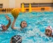 Water polo action shot: a player throws the ball while two opponents swim nearby, creating splashes in the pool.