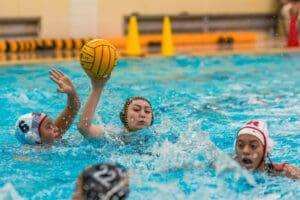 Water polo action shot: a player throws the ball while two opponents swim nearby, creating splashes in the pool.